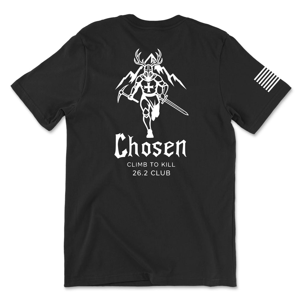 Back view of a black t-shirt featuring the 'Chosen' 26.2 Club graphic in white. The design shows a knight with an antlered helmet holding a sword and a shield with a cross, set against a background of mountain peaks and a sun-arch. Below the warrior, the word 'Chosen' is written in a stylized font, followed by the phrases 'CLIMB TO KILL' and '26.2 CLUB.' A white American flag patch is visible on the right sleeve.