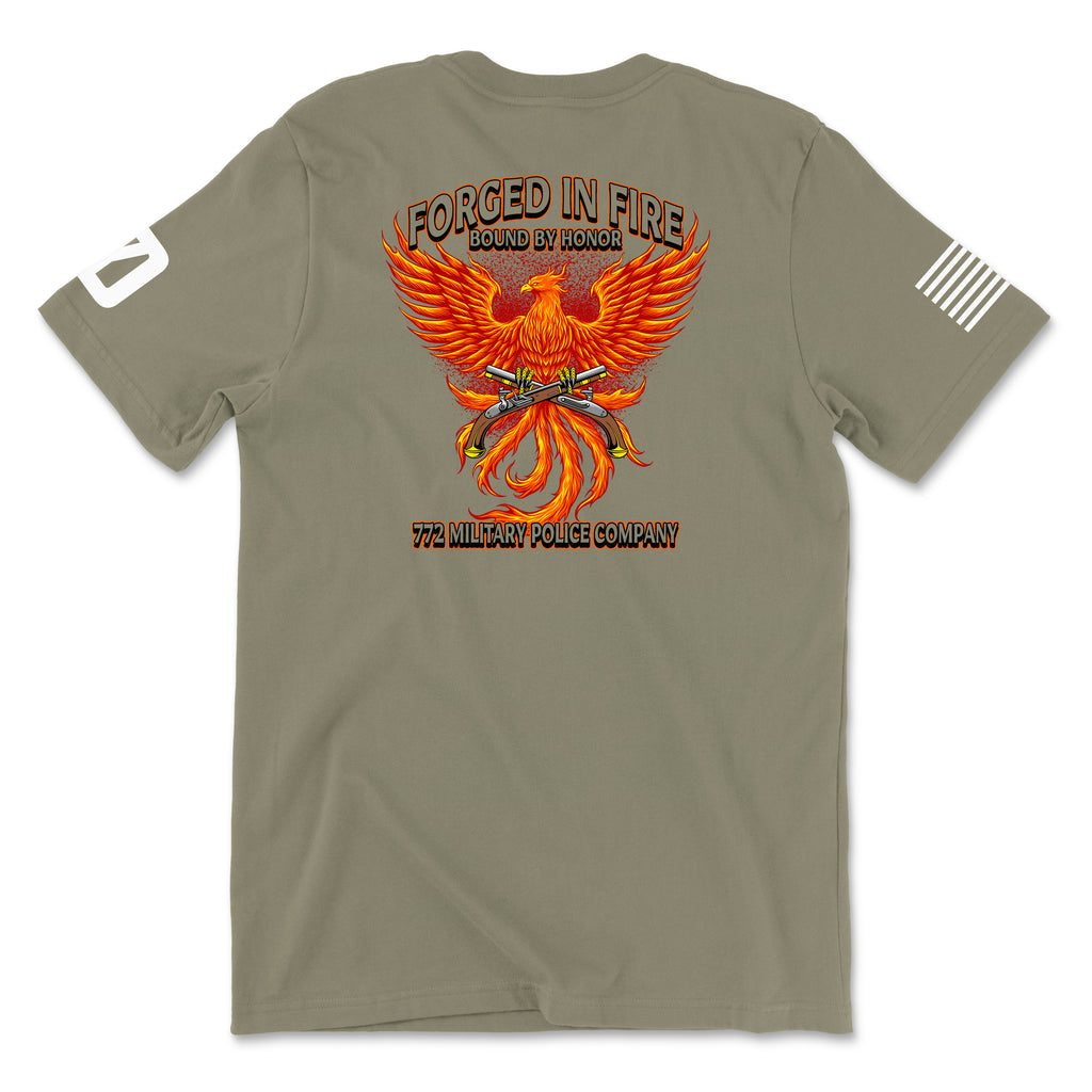 Back view of a coyote brown t-shirt for the 772nd Military Police Company. The design features a large orange and yellow phoenix rising from flames, with crossed flintlock pistols and a blue infantry disc at its center. Banners above read 'FORGED IN FIRE' and 'BOUND BY HONOR,' with '772 MILITARY POLICE COMPANY' written in red at the bottom. A white American flag patch is on the right sleeve.
