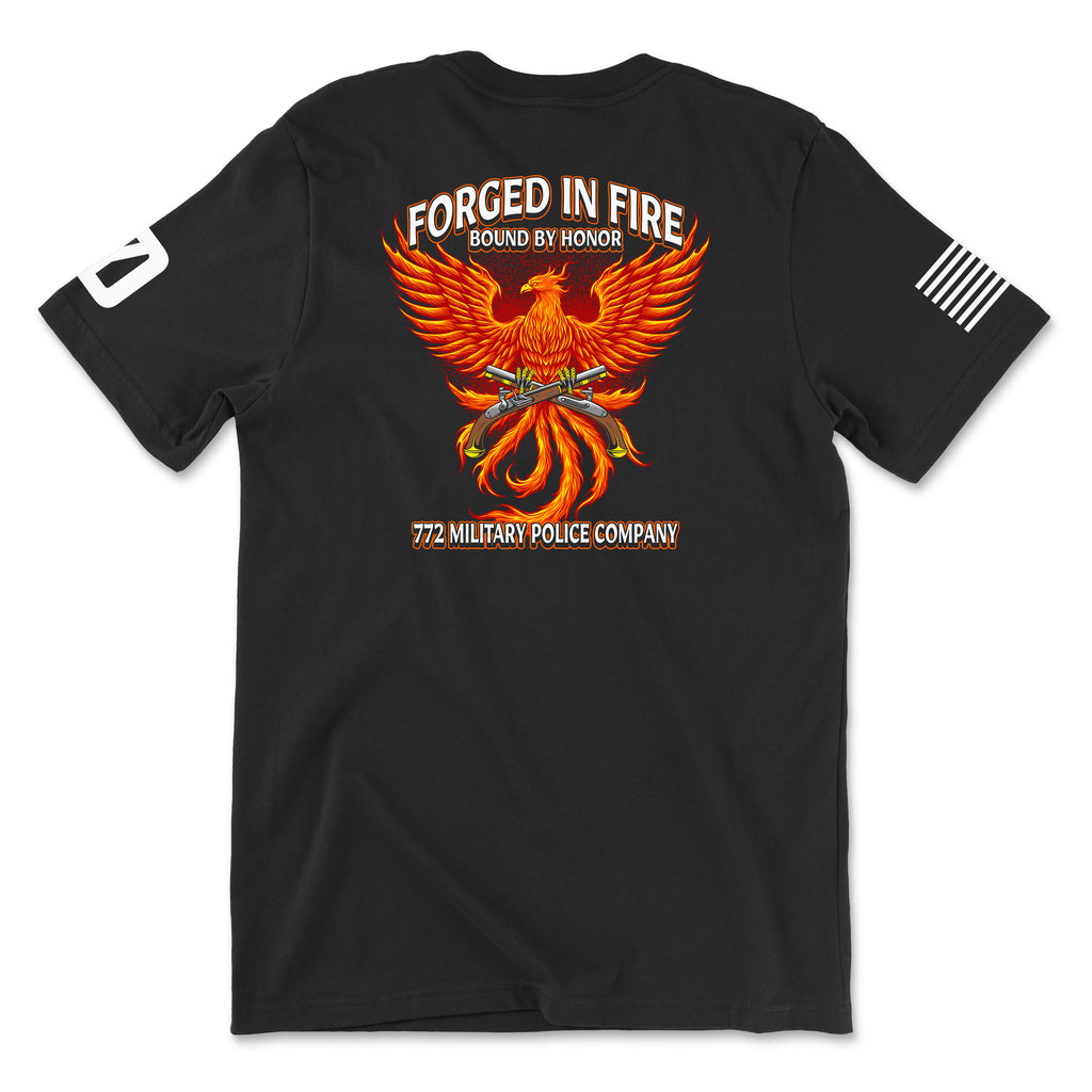 Back view of a black t-shirt for the 772nd Military Police Company. The design features a large orange and yellow phoenix rising from flames, with crossed flintlock pistols and a blue infantry disc at its center. Banners above read 'FORGED IN FIRE' and 'BOUND BY HONOR,' with '772 MILITARY POLICE COMPANY' written in red at the bottom. A white American flag patch is on the right sleeve.