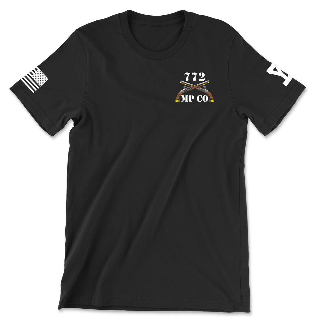 Front view of a black t-shirt for the 772nd Military Police Company. The left chest features a white graphic with the number '772' above a blue infantry disc and crossed flintlock pistols, with 'MP CO' written at the bottom. A white American flag patch is displayed on the right sleeve