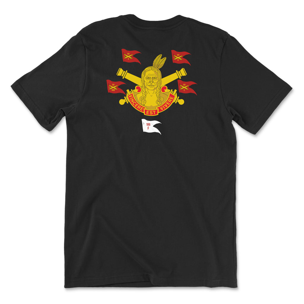 Back view of a black t-shirt featuring the 101st Field Artillery Regiment heritage graphic. The design includes a central figure of a Native American warrior, crossed artillery cannons, and multiple red guidons marked '101.' A red banner at the bottom reads 'VINCERE EST VIVERE' and a small white flag below shows '186 F'.