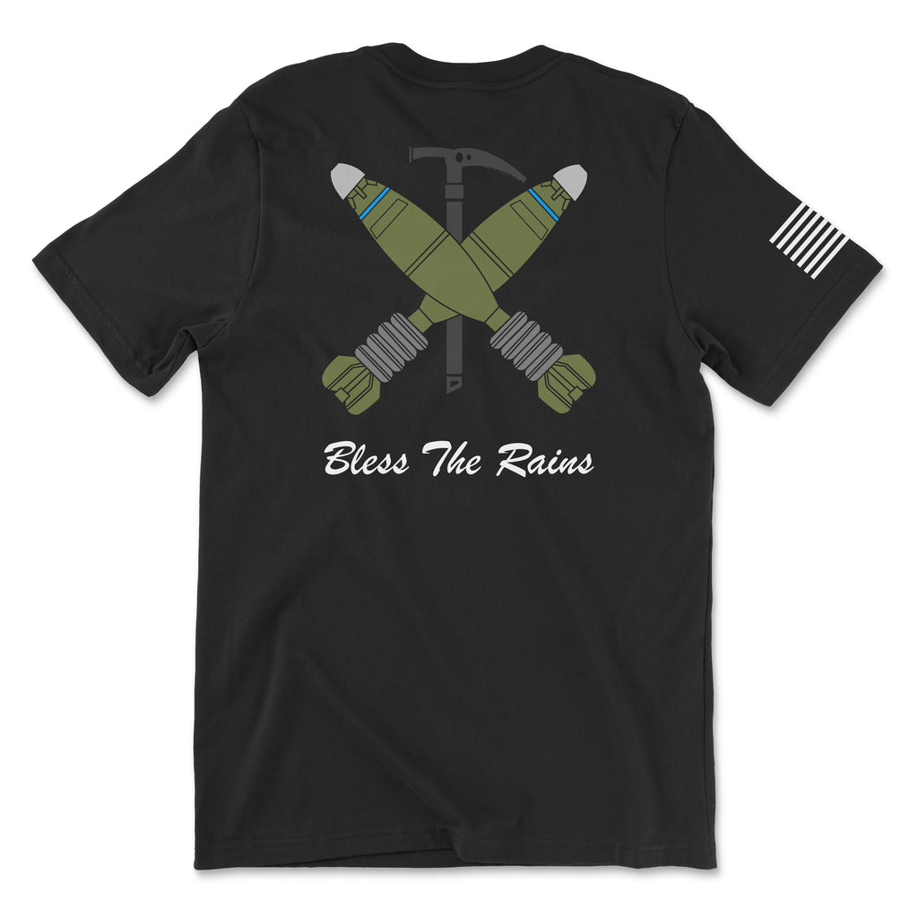 Back view of a black t-shirt featuring a tactical graphic. The design shows two green mortar shells crossed over a black ice axe. Below the graphic, the text 'Bless The Rains' is written in white cursive script. A white American flag patch is visible on the right sleeve.