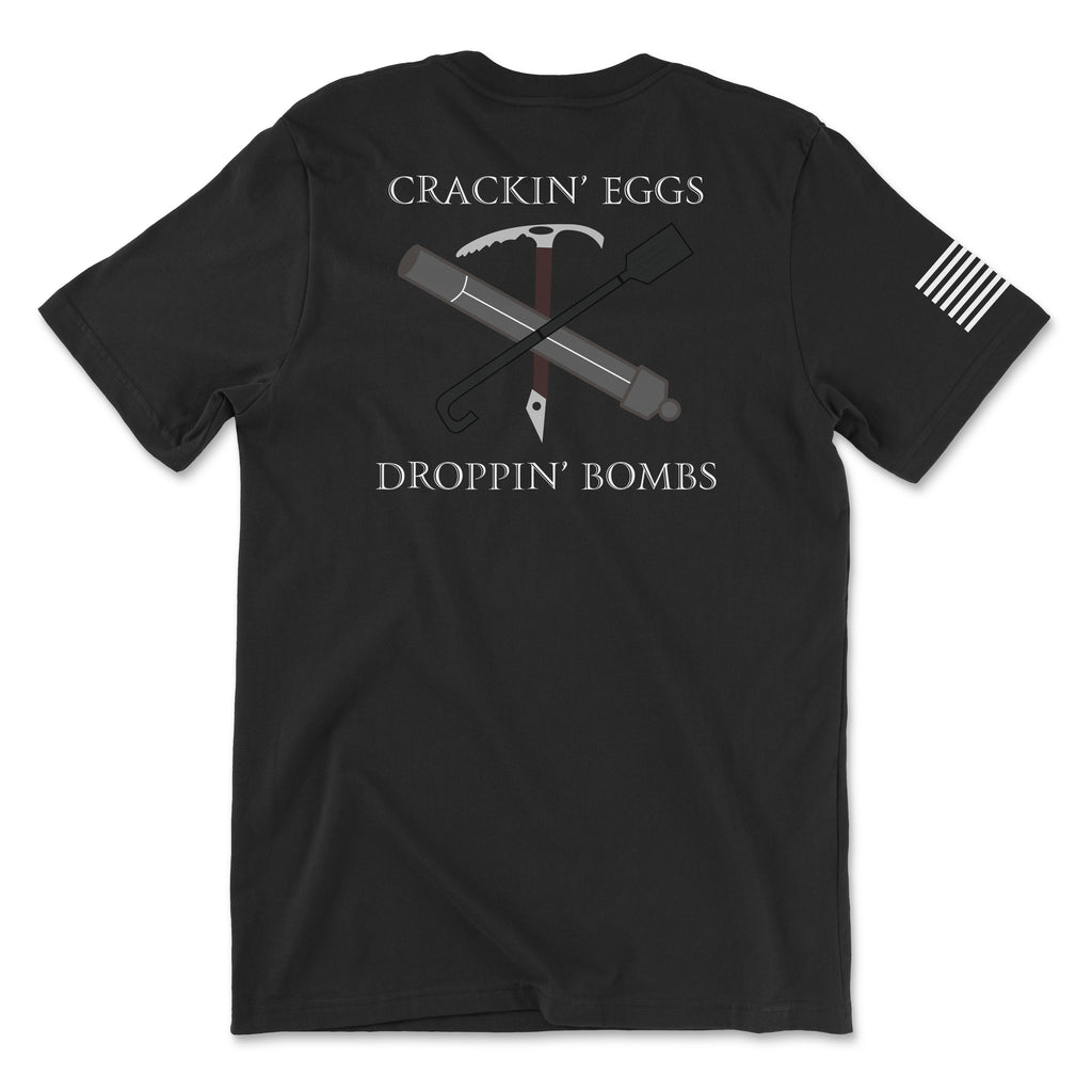 Back view of a black t-shirt featuring the text 'CRACKIN’ EGGS' at the top and 'DROPPIN’ BOMBS' at the bottom in white serif font. The central graphic shows a crossed ice axe and a tactical breaching tool (Halligan bar) with a small white diamond-shaped tag hanging from the center. A white American flag patch is visible on the right sleeve.