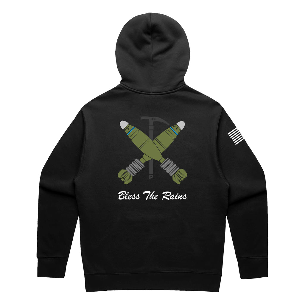 Back view of a black pullover t-shirt featuring a tactical graphic. The design shows two green mortar shells crossed over a black ice axe. Below the graphic, the text 'Bless The Rains' is written in white cursive script. A white American flag patch is visible on the right sleeve.
