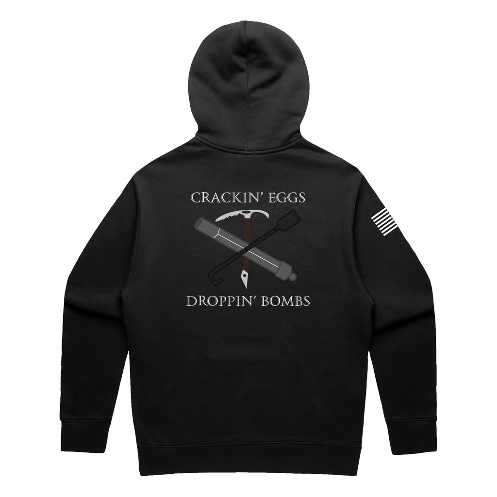 Back view of a black pullover hoodie featuring the text 'CRACKIN’ EGGS' at the top and 'DROPPIN’ BOMBS' at the bottom in white serif font. The central graphic shows a crossed ice axe and a tactical breaching tool (Halligan bar) with a small white diamond-shaped tag hanging from the center. A white American flag patch is visible on the right sleeve.
