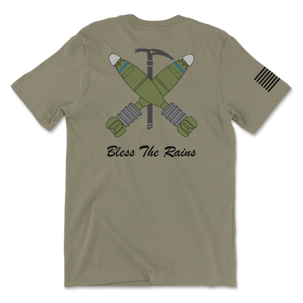 Back view of a coyote brown t-shirt featuring a tactical graphic. The design shows two green mortar shells crossed over a black ice axe. Below the graphic, the text 'Bless The Rains' is written in white cursive script. A white American flag patch is visible on the right sleeve